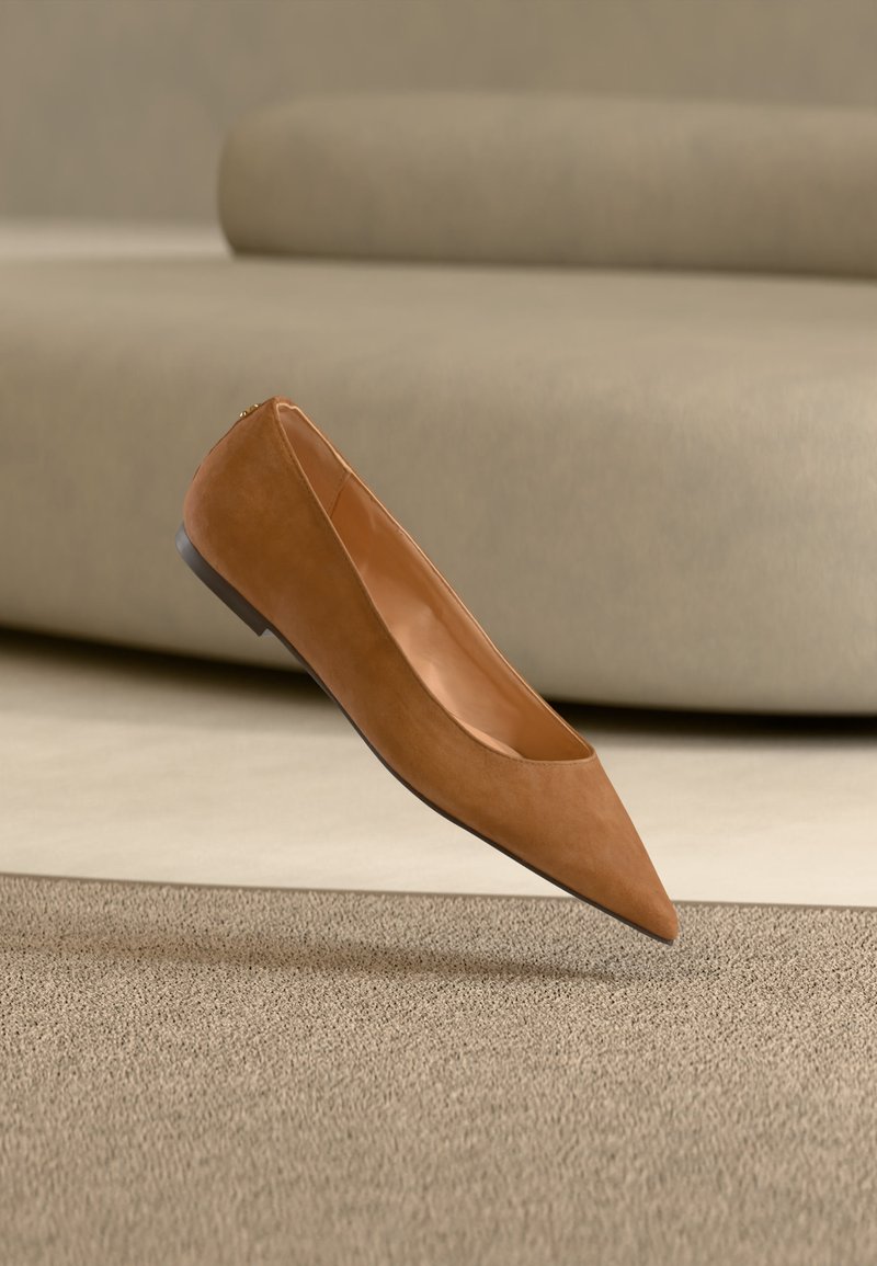 Καφέ επίπεδο παπούτσι μυτερό επιπλέει πάνω από μια υφασμάτινη μπεζ μοκέτα με έναν ανοιχτό μπεζ καμπυλωτό καναπέ στο βάθος.