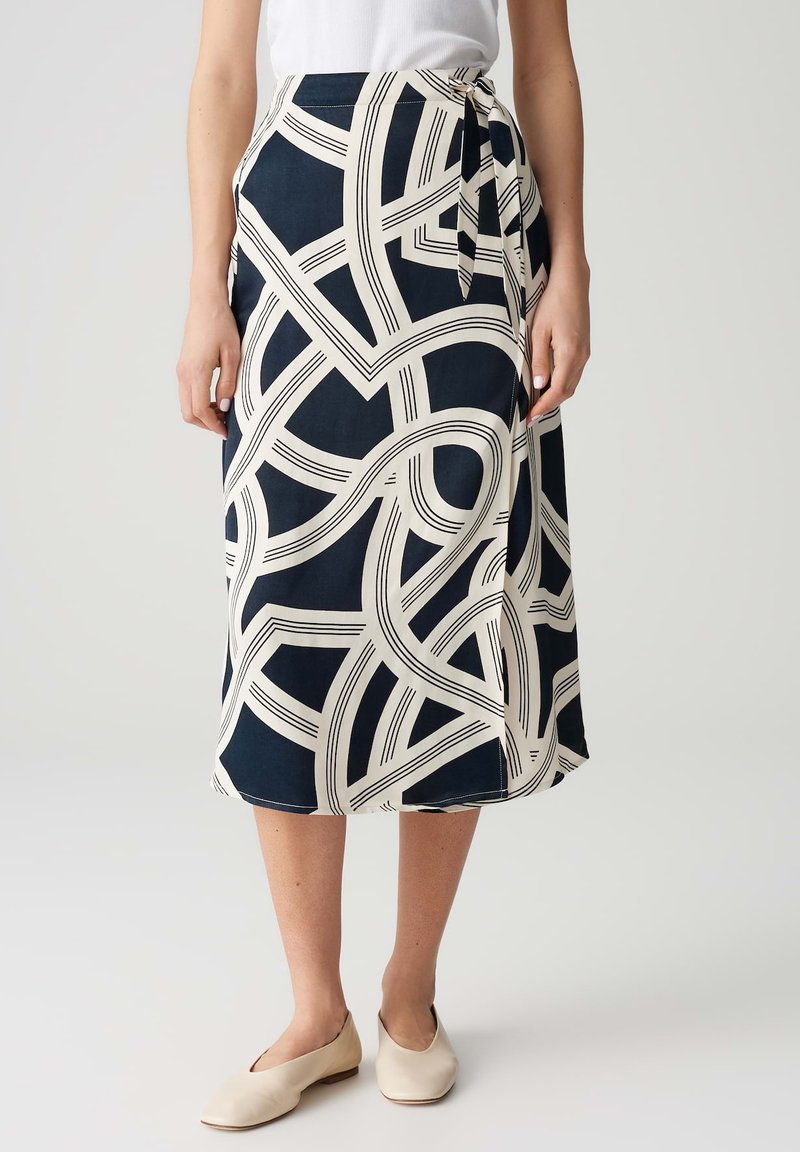 Jupe enveloppante bleu marine avec un motif de rayures abstraites crème, longueur mi-mollet, tissu léger, lien à la taille, complétée par des chaussures claires.