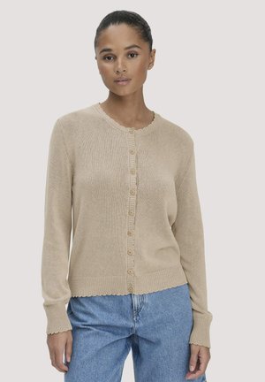 Vrouw die een beige vest met knopen draagt met gegolfde randen en blauwe, hooggetailleerde spijkerbroek tegen een effen achtergrond.