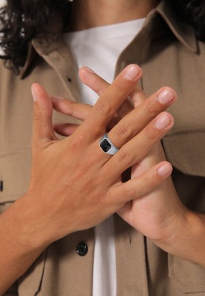 Hõbedast metallist sõrmus ristkülikukujulise musta kiviga, kantud käel, millel on kergelt maniküüriga küüned, pruunil särgil taustal.