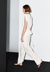 Lichtbeige top met een vloeiend ontwerp en een open rug, gecombineerd met losse beige broek. Model draagt zwarte platte sandalen. Minimalistische setting.