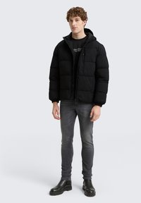 Ung mand iført sort polstret jakke, sort skjorte, grå slim-fit jeans og sorte støvler, stående mod en ensfarvet lys baggrund.