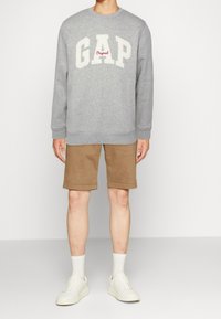 Felpa grigia con la scritta "GAP" e "Original" stampata in bianco e rosso, abbinata a pantaloni corti beige e sneakers bianche, posizionata su uno sfondo neutro.