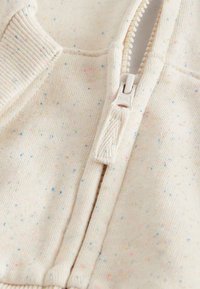 Posnetek blizu kremenastega blaga z mavričnimi pikami, na katerem je delno zadrgana plastična zadrga s tkaninskim trakom za poteg.