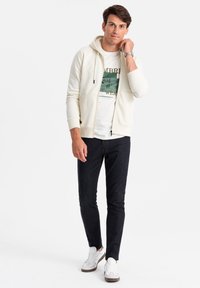 Lichtgekleurde, ritssluiting hoodie met een logo, gedragen over een wit graphic T-shirt. Gecombineerd met donkere jeans en witte sneakers. Eenvoudig en casual ontwerp.