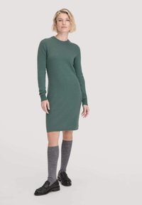 Grünes knielanges Kleid aus weichem Stoff, mit rundem Halsauschnitt und langen Ärmeln. Kombiniert mit grauen Rippensocken und schwarzen Loafers.