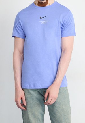 T-shirt en coton violet à manches courtes, arborant un logo Nike noir et un texte gris au centre. Coupe décontractée et texture lisse.