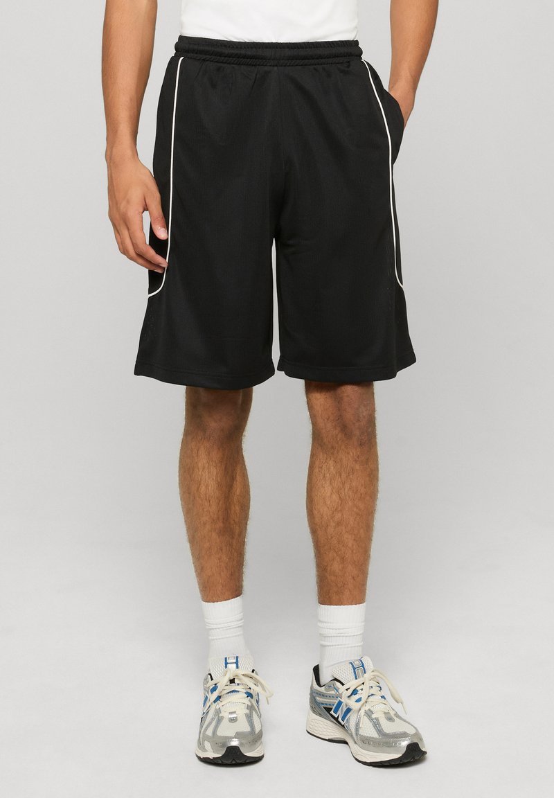 Homme portant un short de sport noir avec des liserés blancs, des chaussettes blanches et des chaussures de course argentées sur un fond clair uni.