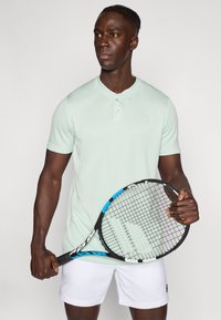 Μέντα πράσινο κοντομάνικο πόλο με υφή, κουμπιά εμπρός και μικρό λογότυπο. Κρατάει ρακέτα τένις με μαύρη λαβή και μπλε λεπτομέρειες.