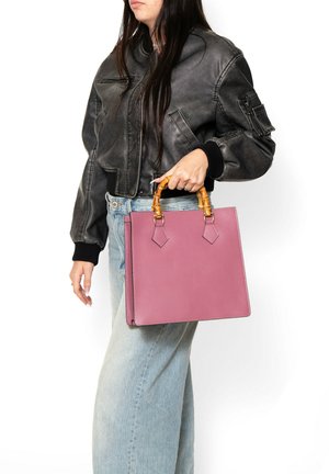 Donna con giacca bomber nera e jeans chiari che tiene una borsa quadrata rosa con manici in stile bambù su sfondo bianco.
