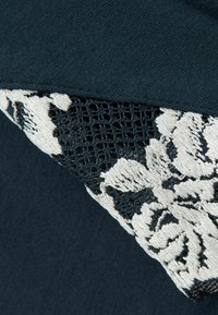 Mörk teal tyg har ett fint spetsöverdrag med svarta och vita blommönster som visar en texturerad kontrast.