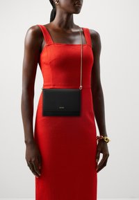 Svart lærcrossbody-veske med teksturert overflate og gulllogo. Tynn metallkjede-stropp. Båret over en tettsittende rød kjole.