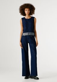 Noor naine lokkis juustega, kes kannab ilma varrukateta tumesinist toppi hõbedaste servadega, sobivaid laiad pluusid ja musti mokasiine, seisab valge tausta ees.