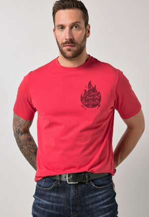 Uomo con barba e braccio tatuato che indossa una maglietta rossa con grafica di fiamma nera e testo, abbinata a jeans blu e cintura nera, in piedi con le mani dietro.