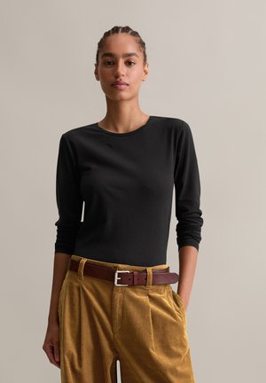 Woman with braided hair wearing a black long-sleeve shirt tucked into mustard corduroy pants with a brown leather belt.