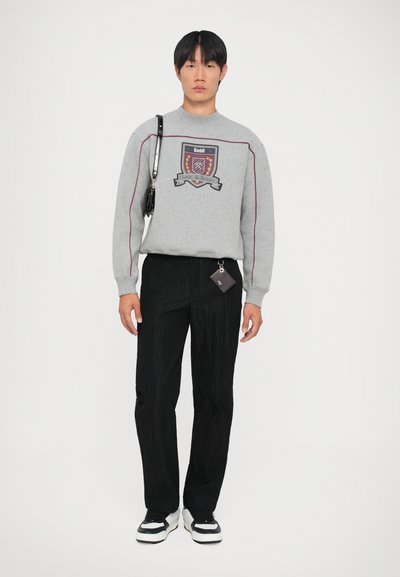 Felpa grigia con grafica a scudo, pantaloni neri texturizzati e sneaker bianche con dettagli neri, accompagnati da una piccola borsa nera.