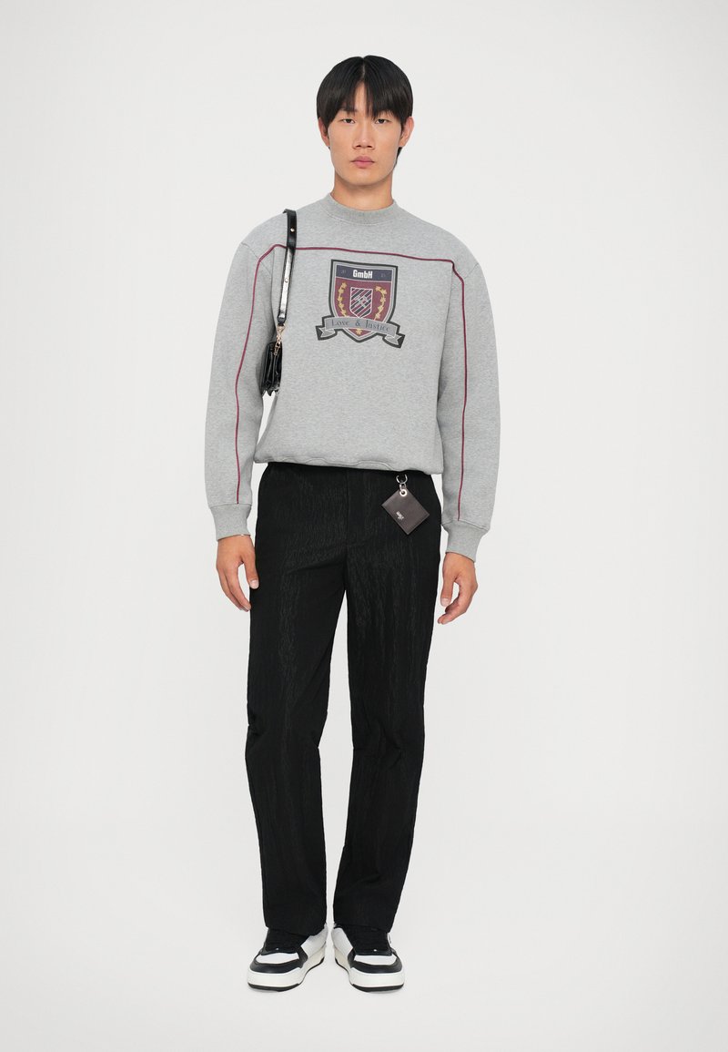 Felpa grigia con grafica a scudo, pantaloni neri texturizzati e sneaker bianche con dettagli neri, accompagnati da una piccola borsa nera.