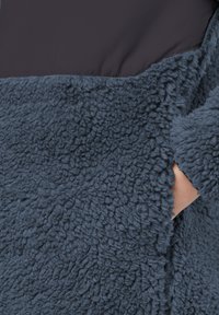 Couche extérieure en polaire de couleur bleu foncé avec une surface texturée et duveteuse. Tissu lisse en haut dans une couleur contrastante. Poche latérale visible.