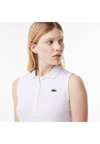 Polo shirt bianco senza maniche con colletto, caratterizzato da un logo di coccodrillo ricamato di colore verde sul lato sinistro del petto. Texture liscia.