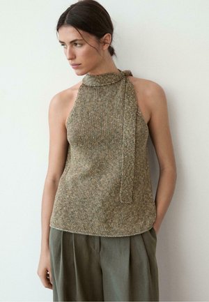 Femme aux cheveux foncés attachés, portant un haut sans manches en tricot beige style licou et un pantalon ample vert olive, regardant vers la gauche.