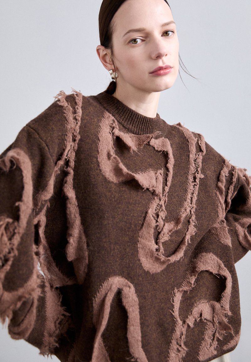 Bruine trui met rafelige, textuurpatches in een lichter bruin ontwerp. Heeft een geribbelde halslijn en een oversized pasvorm.