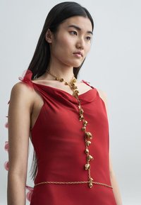 Vörös, ujjatlan ruha, drapériás nyakkivágással, áttetsző rózsaszín díszítéssel a vállon és egy aranylánccal, levél alakú medálokkal.