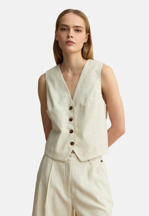 Mujer con chaleco sin mangas beige con botones y pantalones de talle alto a juego, de pie con las manos detrás de la espalda.