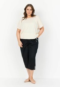 Beige kortärmad topp i kombination med svarta cropped-byxor med sidofickor och knappdetaljer. Modellen står med en hand på höften.