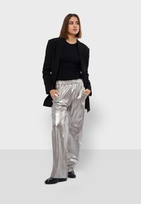 Zilver metallic wijde broek met elastische tailleband, gecombineerd met een zwarte fitted top en een zwarte blazer, staand met de handen in de zakken.