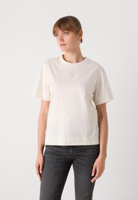 T-shirt di colore crema, a maniche corte, realizzata in tessuto morbido, con un piccolo logo ricamato sul petto. Taglio dritto con un orlo leggermente più lungo.