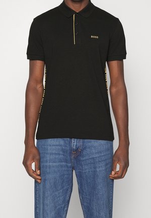 Homme portant un polo noir BOSS avec logo et bordure jaunes, associé à un jean bleu, debout devant un fond uni.