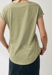 Kortærmet, olivengrøn t-shirt med en rund søm og opfoldede ærmer, lavet af et blødt materiale. Båret med en mørk denimnederdel.
