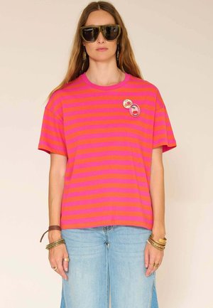 Femme portant un t-shirt oversize rayé rose et orange avec des pins fruités, un jean bleu clair, de grandes lunettes de soleil et des bracelets.