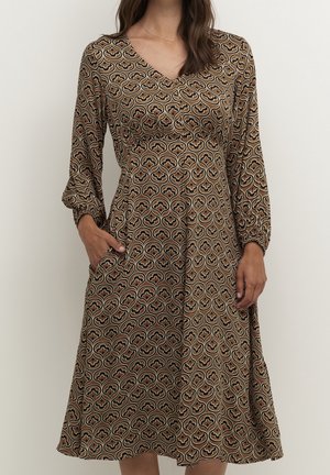 Kobieta w brązowej, wzorzystej sukience midi z długimi rękawami, dekoltem w serek i bocznymi kieszeniami, stojąca na jednolitym jasnym tle.