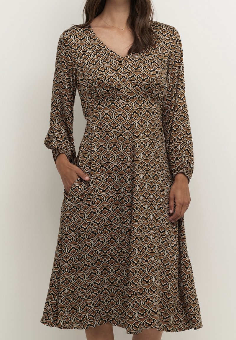 Femme portant une robe midi marron à motifs avec des manches longues, un col en V et des poches latérales, se tenant devant un fond clair uni.