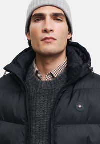 Sort pufferjakke med høj krave, prydet med et logo-patch. Båret over en grå strikket sweater og en brun ternet skjorte, med en grå hue.