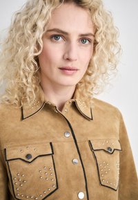Camicia in suede beige con borchiette argentate, cuciture a contrasto nere e due tasche sul petto. Presenta un colletto classico e bottoni a pressione.