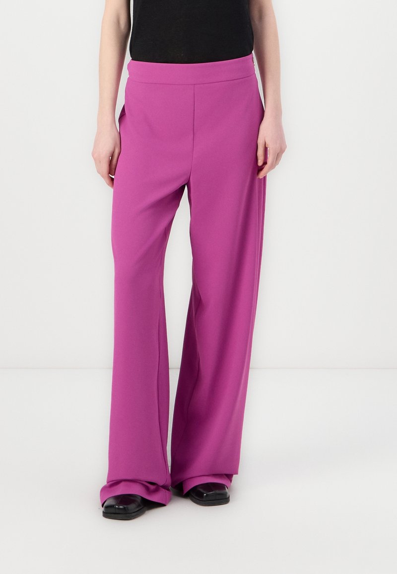 Femme portant un pantalon taille haute, jambes larges de couleur magenta, associé à des chaussures noires à bout carré et un haut noir, debout devant un fond uni.
