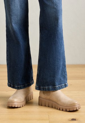 Beige ankelstøvler med en teksturert overflate og kraftige, rillete såler. Kombinert med mørkeblå flagrende jeans og satt mot et tregulv.