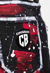 Denim noir avec éclaboussures de peinture rouge, blanche et grise ; présente un patch logo noir "CIPO&BAXX" et des surpiqûres blanches. Design à cinq poches.