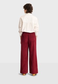 Pantaloni in velluto a coste rossi a gamba larga con tasche posteriori, abbinati a una camicia bianca con bottoni. Texture morbida con una vestibilità rilassata.