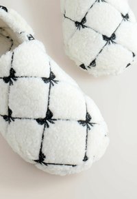 Chaussons en peluche blancs avec un design matelassé, ornés de nœuds noirs et d'une texture douce. Bout rond et doublure douce pour le confort.