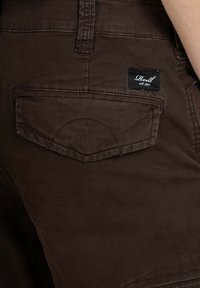 Braune Baumwoll-Cargo-Shorts mit einer Gesäßtasche, die einen gewölbten Klappenverschluss hat, und einem schwarzen Etikett mit weißem Text, das angenäht ist.