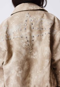 Veste en daim beige avec des clous métalliques disposés en forme d'étoile et un texte en cursive partiellement visible au dos.