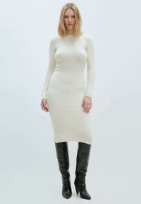 Strick-Midi-Kleid in Creme mit gerippter Textur, langen Ärmeln und einer taillierten Silhouette. Kombiniert mit glänzenden schwarzen kniehohen Stiefeln.