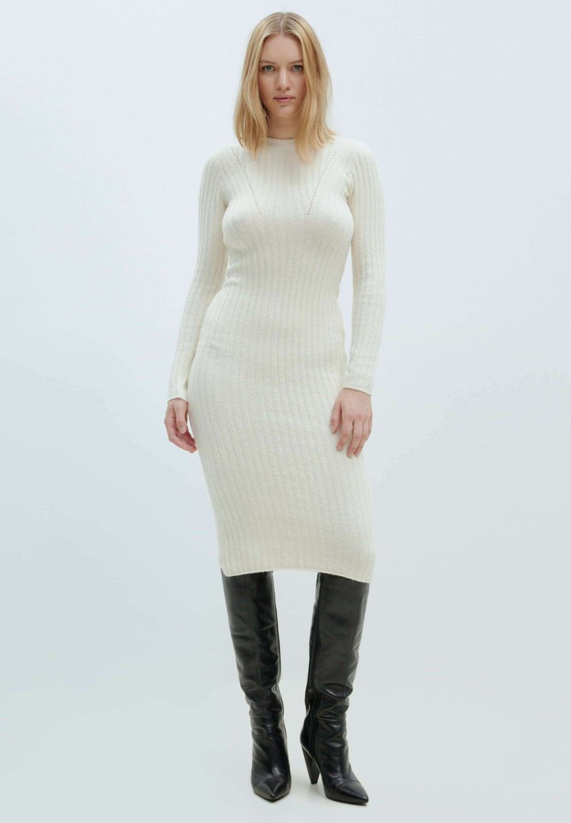 Strick-Midi-Kleid in Creme mit gerippter Textur, langen Ärmeln und einer taillierten Silhouette. Kombiniert mit glänzenden schwarzen kniehohen Stiefeln.