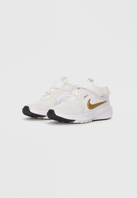 Para białych sneakersów Nike z złotym logo Swoosh, rzepami i czarnymi podeszwami na jednolitym białym tle.