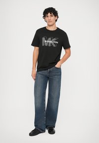 Mladý muž s kučeravými vlasmi, oblečený v čiernom tričku s logom Michael Kors, modrých širokých džínoch a čiernych teniskách, stojí so jednou rukou v vrecku.