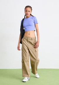 Lila crop t-paita, jossa on logo, yhdistettynä beigeihin leveisiin cargo-housuihin, joissa on sivutaskut. Asusteena musta laukku ja valkoiset tennarit.