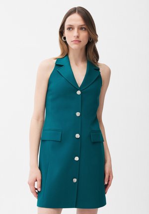 Kobieta ubrana w bezrękawnikową, morsko-zieloną sukienkę z kołnierzem z klapą i pięcioma srebrnymi guzikami z przodu, stojąca na tle jednolitego, białego tła.